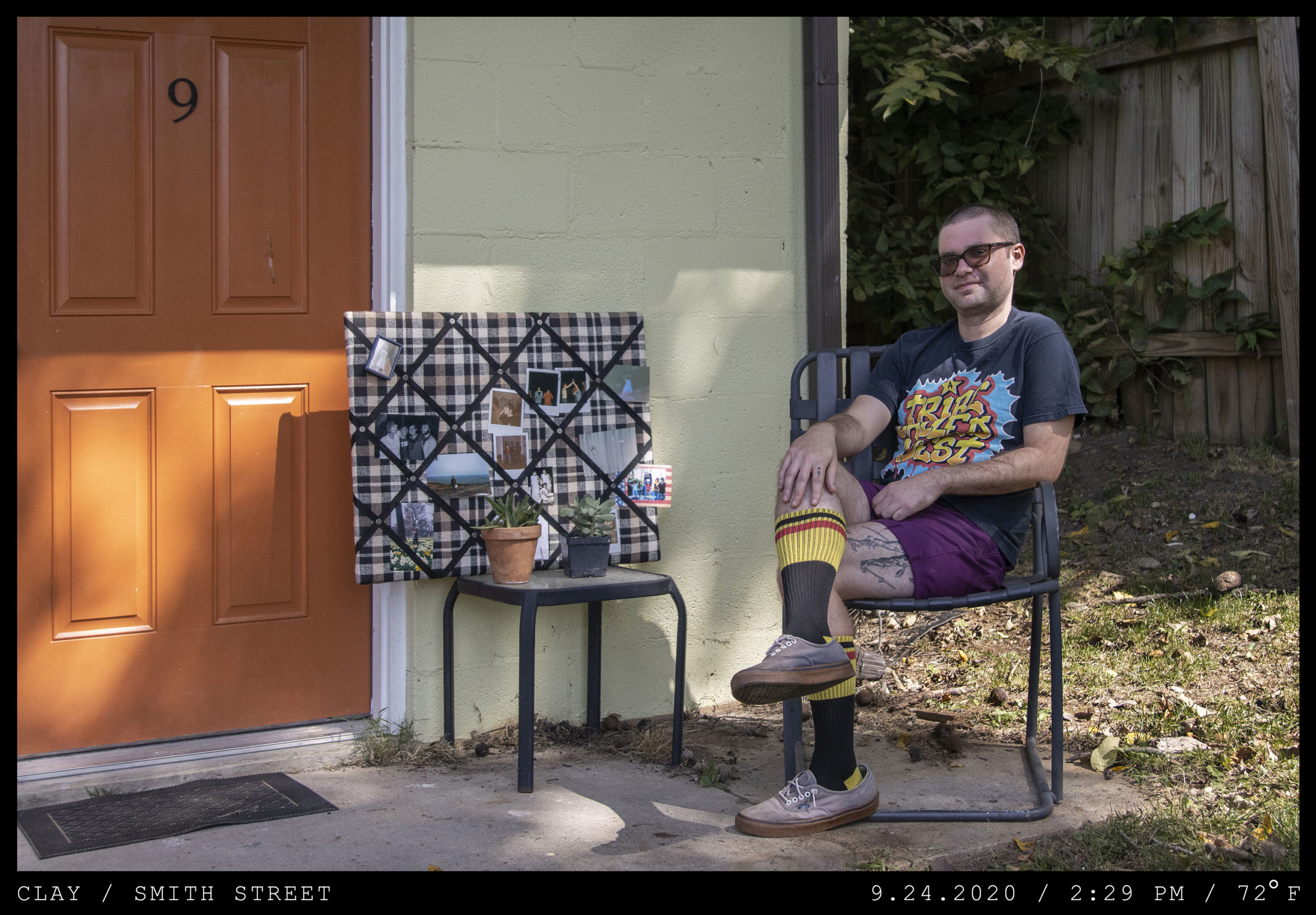 A young man in sunglasses is seated on a front porch in a colorful tee shirt and high yellow socks.