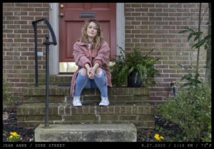 A woman with long wavy hair in distressed jeans and white high tops is seated on a brick stoop.