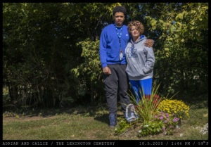 A man and woman stand closely together wearing University of Kentucky athletic wear next to a grave decorated with flowers.