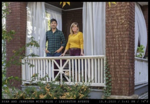 A man with shortm unkempt hair stands in a green and blue flannel shirt next to a blonde woman in a thin yellow sweater on a porch while a tan dog peeking through the rails.