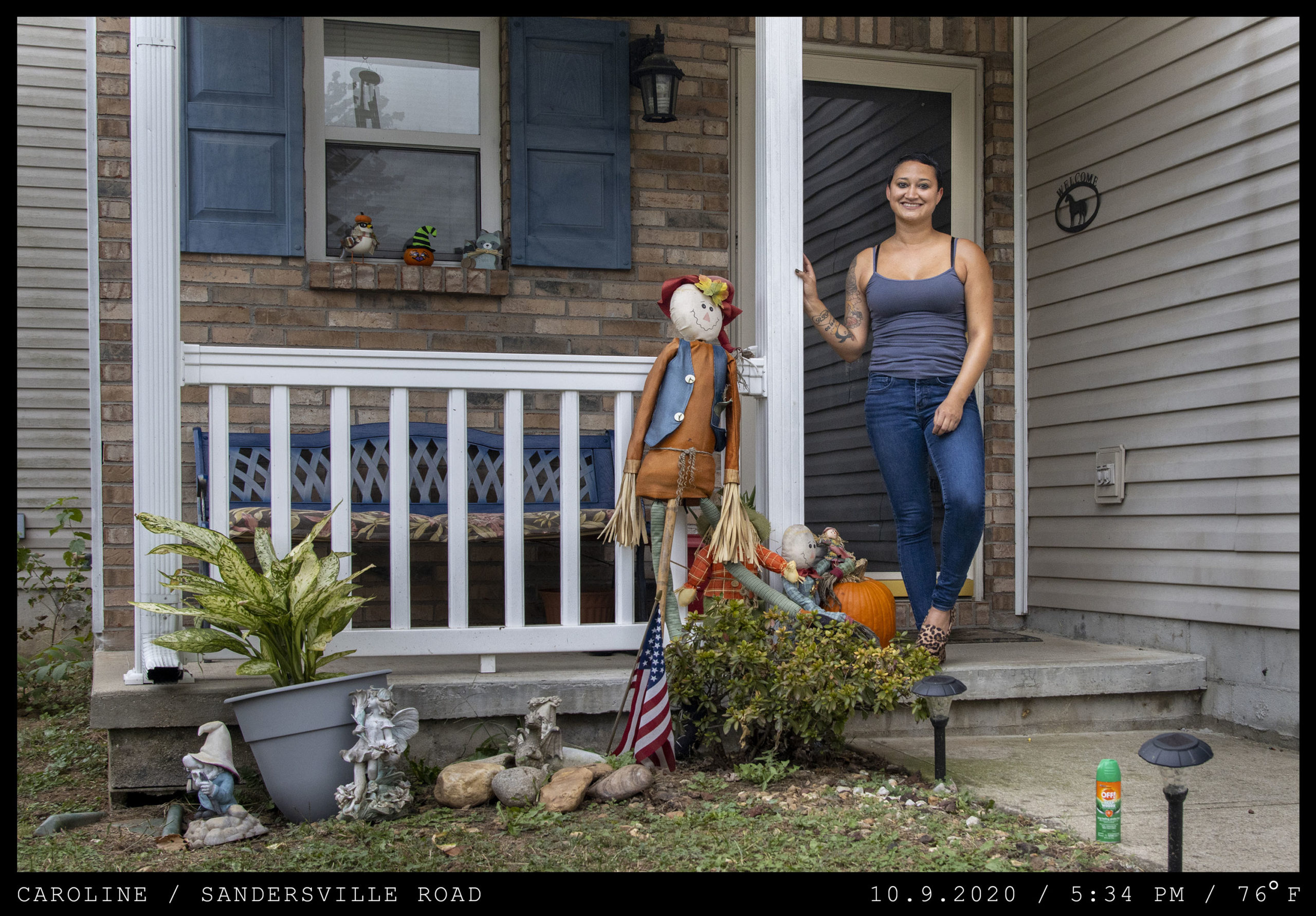 A woman in a gray tank top stands on a front porch featuring Fall decorations and a small American flag.