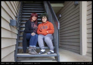 Two women are seated on the stairs of an apartment complete. One wears glasses with dyed dark-auburn hair and a long black cardigan and the other has curly green hair, her hands in the pockets of an orange GAP hoodie, wearing white socks and athletic sandals.