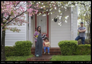 A woman with dark-rimmed glasses and long brown hair wears a floral sweater while standing next to a jumping toddler in a tie-dyed tee shirt. A cross-armed man stands behind shrubs to their left in front of a white, wood panelled home and a brick stoop.