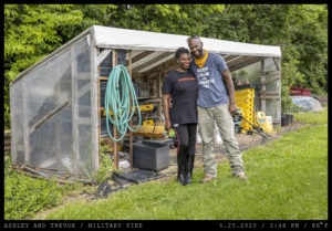 A couple: (left) a young woman with a long tee shirt and rubber boots, (right) a bald man with a beard, sunglasses propped on his forehead, and a yellow handkerchief around his neck, stand in front of an open-front shed filled with gardening supplies and tools.