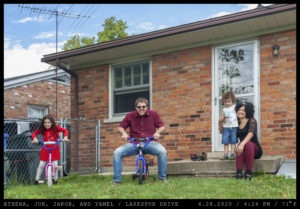 A young family enjoys their back yard: a girl in a red dress on a bicyle and a man in a marron button down are both seated on children's bicyles donning a snarling expression. The man wears sunglasses while a woman in a dark shirt with laced sleeves and curly hair rests her arm around a toddler with short curly hair drinking juice.