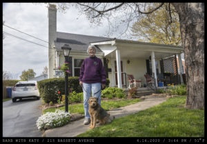 An smiling elderly woman holds an old dog on a leash next to the driveway of a white home with a front porch.