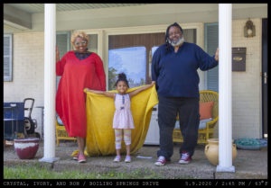 A woman in a bright red dress and curly lightened hair with thick glasses stands next to a young girl with curly dark hair donning a yellow blanket as a cape (arms stretched wide) while a man with dreadlocks and a beard wearing red sneakers leans on a porch column.