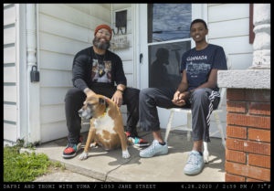Two men, (left) wearing a red woven cap and a black graphic tee shirt and (right) a Granada tee-shirt sit on a front stoop petting an old, brown dog with a colorful collar.