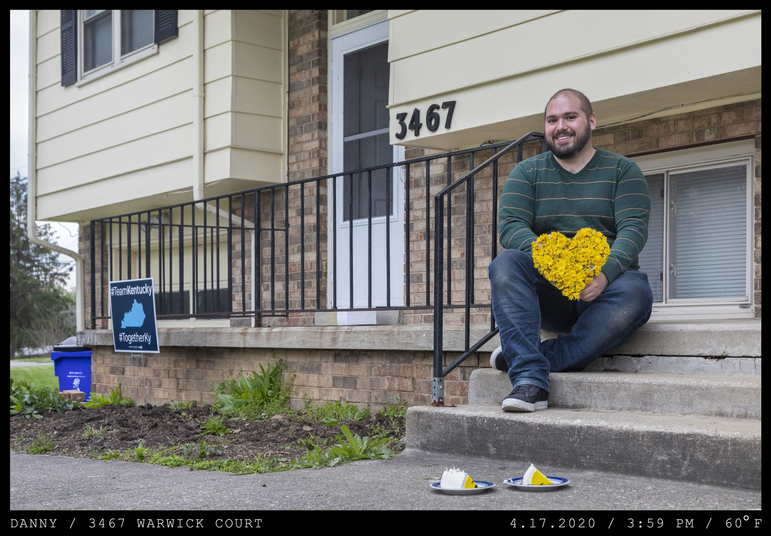 A man in a green striped shirt and jeans with a crew cut and beard is seated on concrete steps holding a yellow floral arrangement in the shape of a heart. Two slices of cake are at his feet on plates.