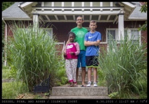 A middle aged man with round glasses stands in front of a brick home with two children, (left) a young girl in braids and pink pajamas and a boy (right) in a blue tee shirt with his arms crossed.