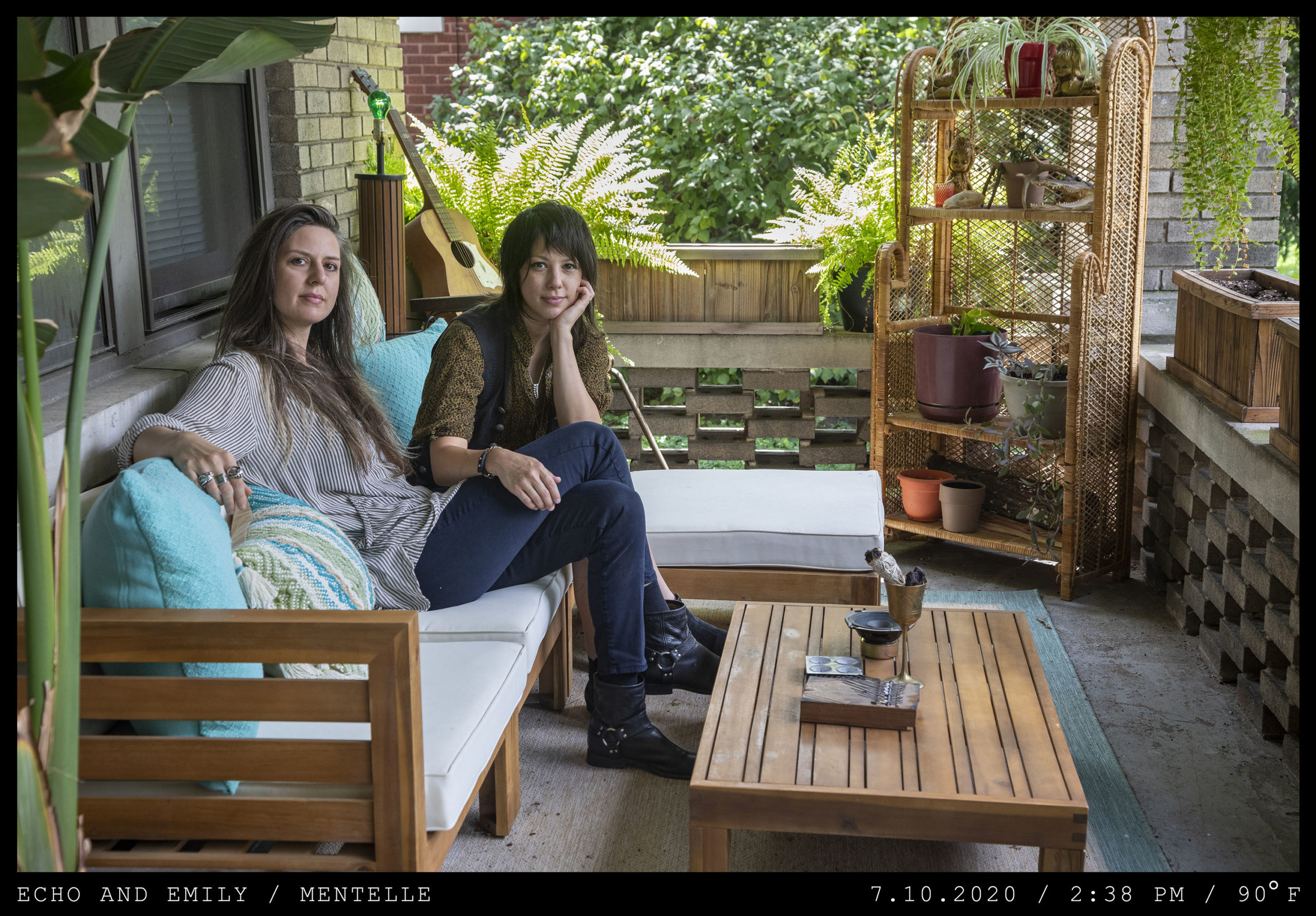 Two women with long dark hair, one with bangs are seated on a front porch among wooden furniture. One sits with legs crossed while the other drapes her right hand across her knee.
