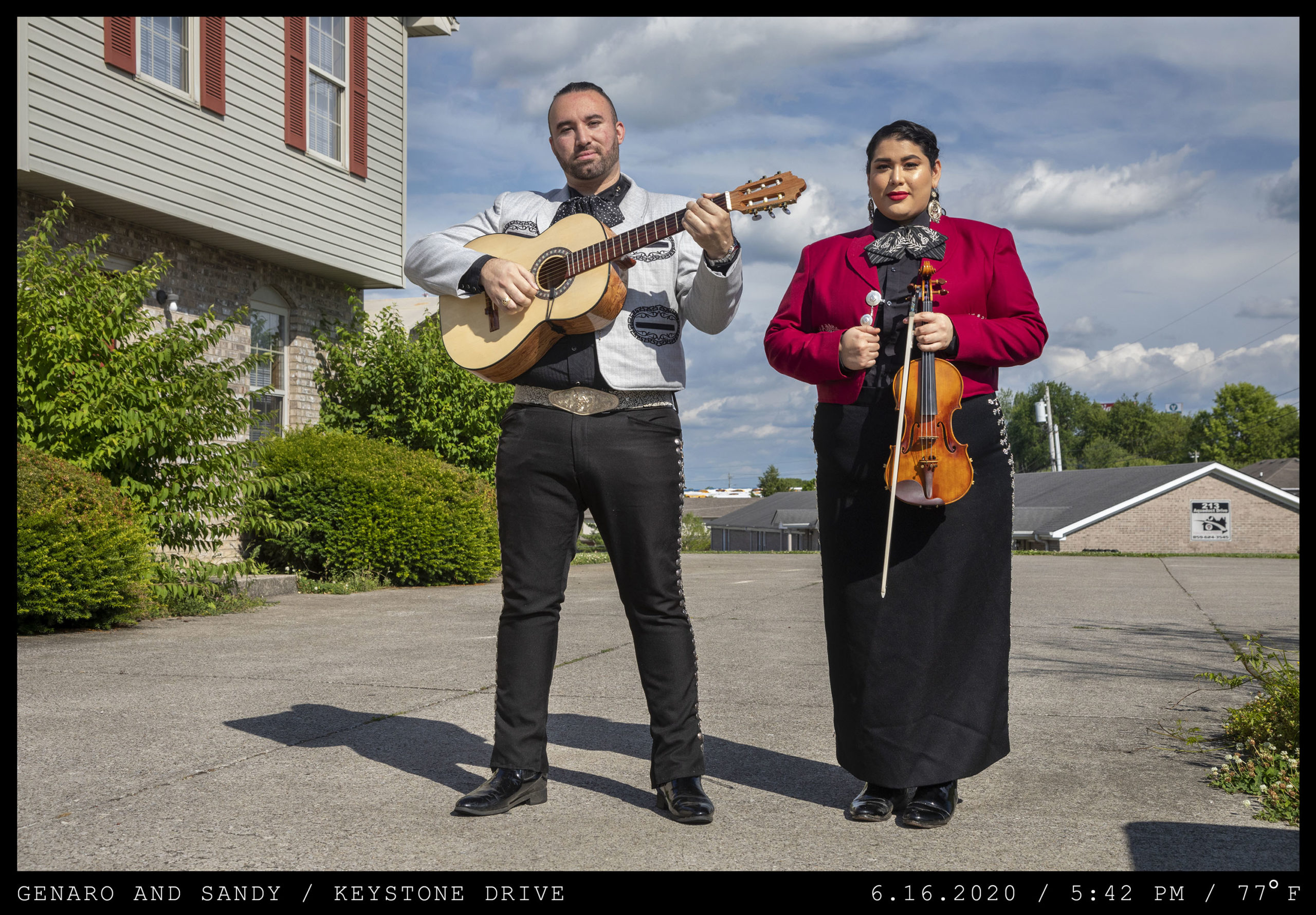 A man with a guitar and woman with a violin stand in neatly pressed Mariachi garb on the concrete driveway of an apartment complex.