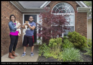 Two people hold cats: a woman with a neon pink shirt and a denim jacket stands next to a man in a Coors tee shirt at the front entryway of a brick home with a white framed arched window.