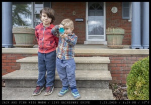 Two young boys stand on the concrete steps of a brick home with blue columns; one (left) has his arms crossed behind his back while the other in colorful gingham tilts a blue cup open revealing an earthworm.