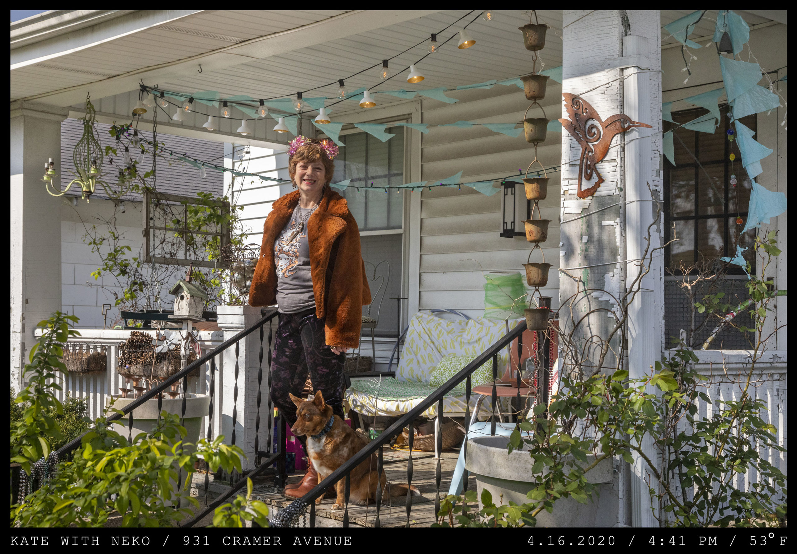 A woman wearing a red and white bow stands on a front porch with patio decorations, plants and chimes. She is wearing a thick, furry rust-colored collared coat in pajama pants with a dog at her side looking down the stairs.