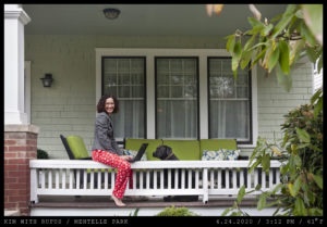 A woman with parted curly hair in a business jacket and red pajama pants with a snowflake pattern is seated with a laptop on the rail of a front porch while a black labrador retriever looks on.
