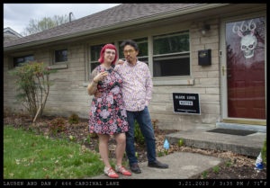 A woman with bangs, pink hair and retro glasses in a floral pink knee-length dress holds a cocktails with her hand on the shoulder of a man in glasses, Van Dyke style facial hair in a colorful polka-dot button up and jeans stand in front of a stone home with a red door donning a skeleton behind a glass storm door with the home number "666." A Black Lives Matter yard sign is left of the stoop.