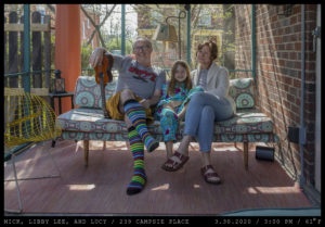 A man holds a ukelele upside-down in colorful, knee-length socks crossed legged seated next to a young girl in brightly patterned pajamas next to a woman in a tan collared sweater and short hair on a patterned coushined bench in a screened porch with orange columns and teal trim.