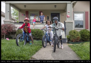 A family of five stand afront a tan home with a columned porch. Three children ride bicycles (a girl in pigtails and two boys wearing helmets), one in a pink shirt is seated on the porch railing while a woman stands beside her.