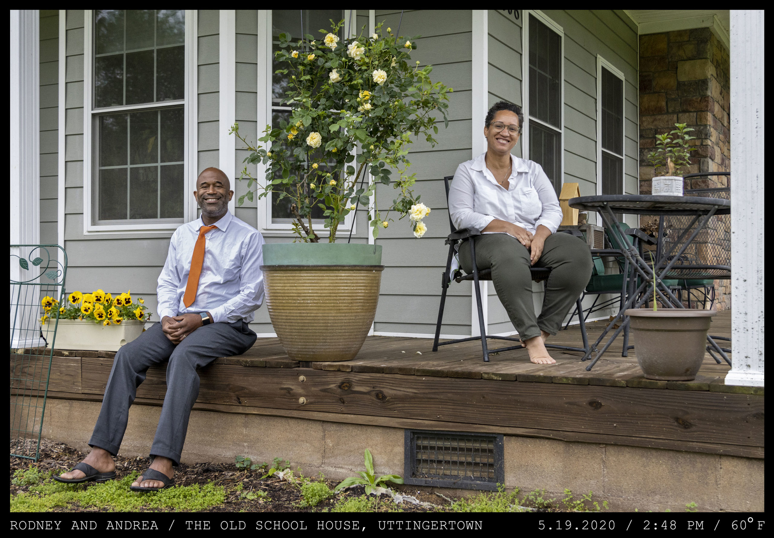 A neatly dressed bald man in an orange tie and leather sandals is seated on the wooden porch of a light freen home next to a woman with short curly hair, barefoot wearing a white button up shirt.
