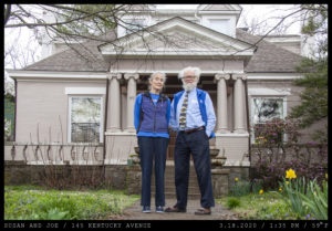 An elderly couple stand in front of a cream brick home with ornate columns. They both wear blue athletic sweater vests. The woman had glasses hanging from a chain on her neck. The main has unkempt white hair, a beard, a blotted patterned tie, and Birkenstocks.