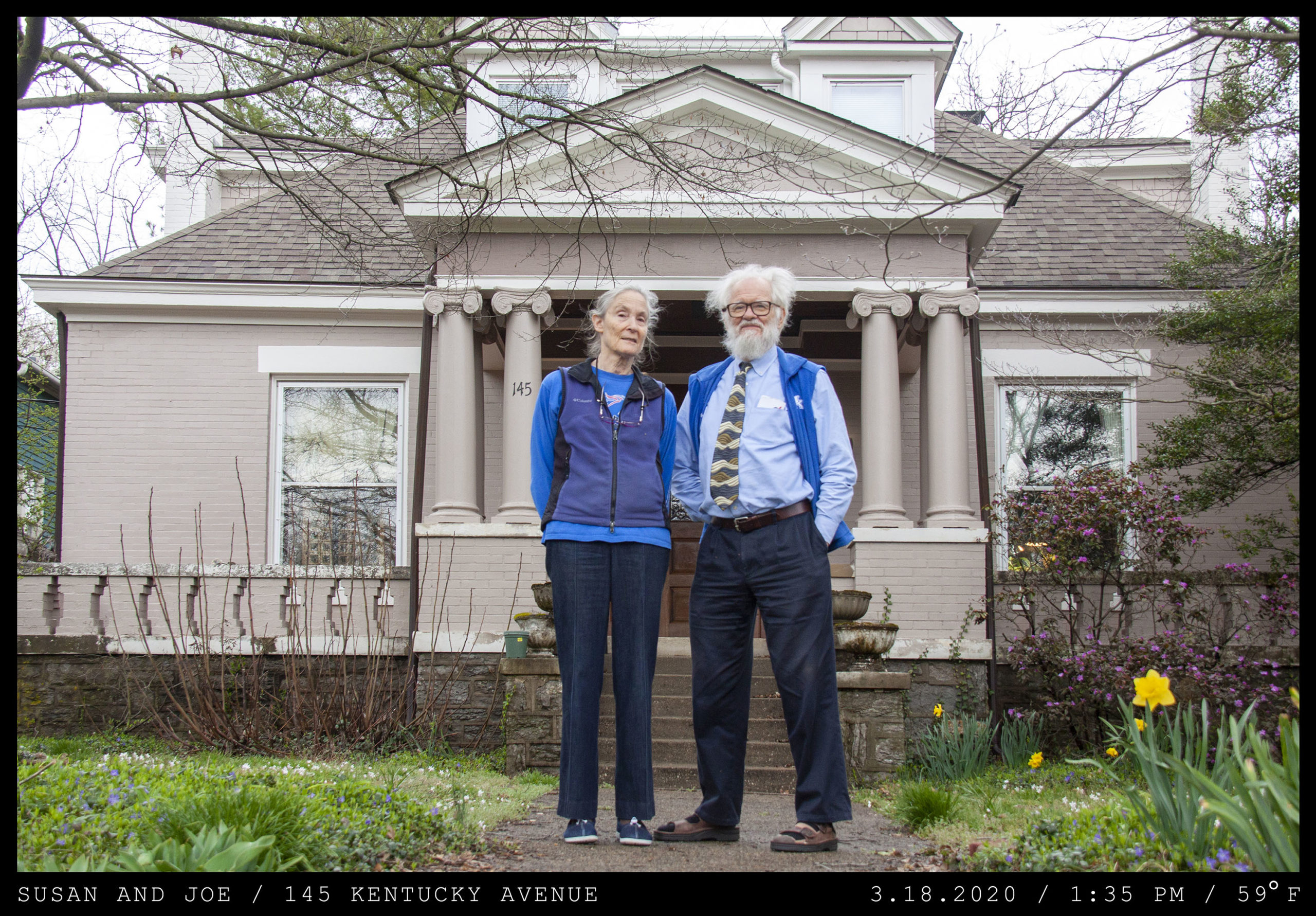 An elderly couple stand in front of a cream brick home with ornate columns. They both wear blue athletic sweater vests. The woman had glasses hanging from a chain on her neck. The main has unkempt white hair, a beard, a blotted patterned tie, and Birkenstocks.