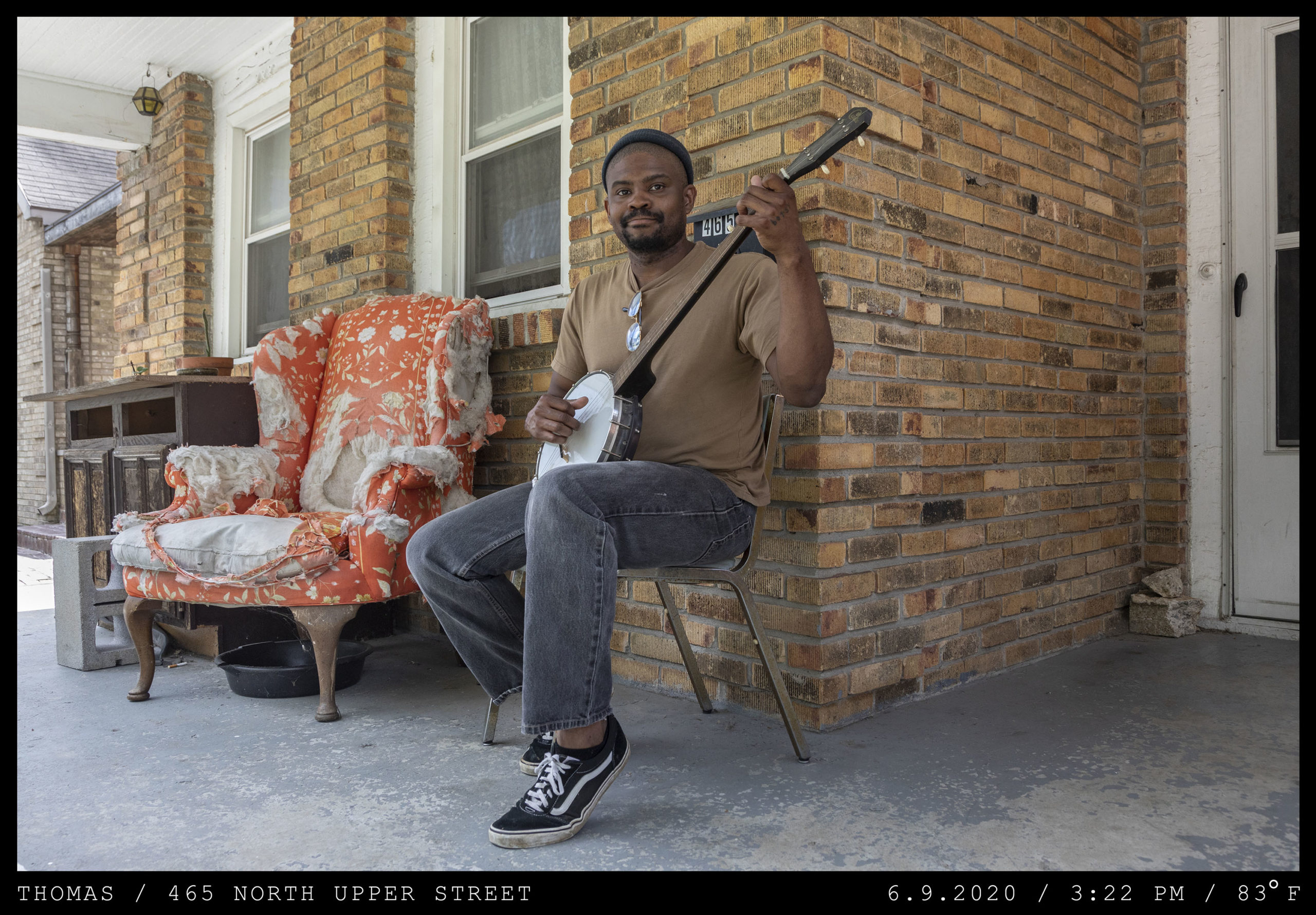 A young man with a beard and cap is seated on the front porch of a brick duplex holding a banjo seated next to a well worn orange upholstered chair.