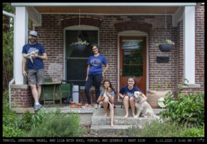 A family of four: a bearded man holding a cat in a light cap leans casually, a woman with short curly hair, and two girls seated on concrete steps stand on the porch of a brick home.