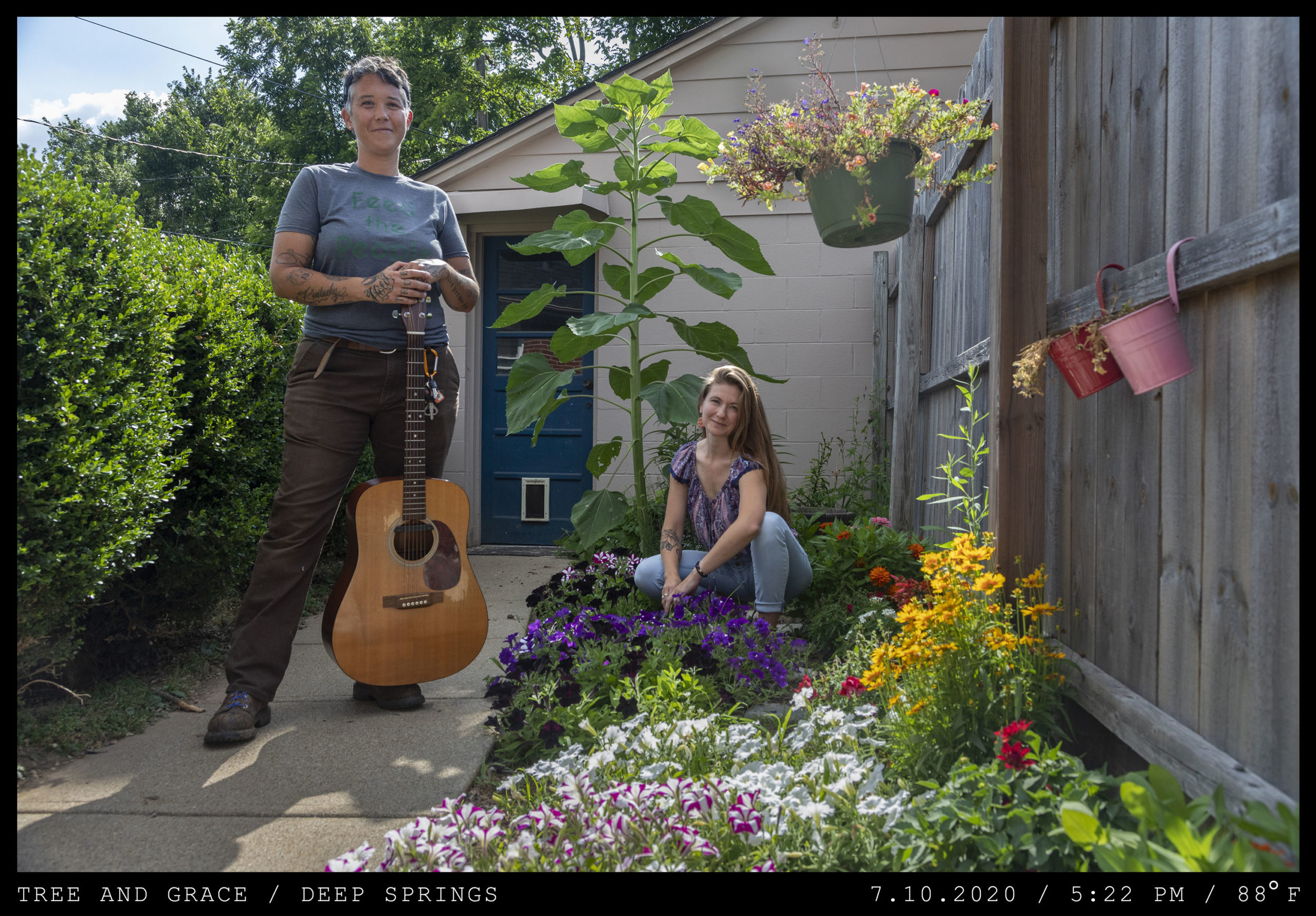 A tattooed young woman with short hair stands in a garden holding an acoustic guitar while a young woman with long brown hair crouches among purple flowers.