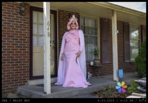 A lavishly dressed person in drag queen attire (a pink dress, cape, tiara, and detailed make up) stands on the concrete stoop of a brick home.