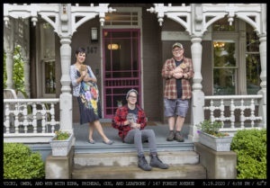 A woman in a floral dress with short hair leans on a column next to a seated young man in glasses wearing a woven cap while an older man in orange flannel, cargo shorts and a cap all stand in front of a VIctorian style home on the front porch holding cats while another looks on from inside the pink framed screen door.
